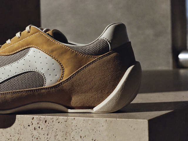 Detaliu macro al unui sneaker: suede nisipiu periat, piele netedă alb-os, plasă fină gri și cusături duble tonale deasupra unei tălpi modelate, pe travertin poros.