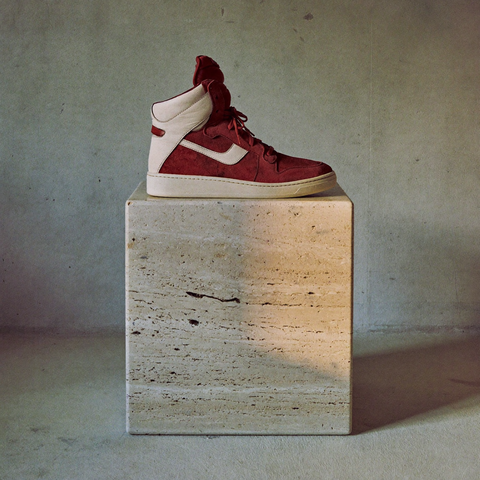 Sneaker înalt din suede vișiniu cu detalii din piele alb-os, în profil, pe un soclu cub de travertin.
