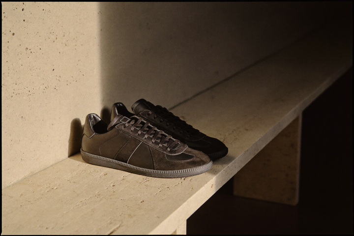 Pereche de sneakers joși din suede negru, pe marginea unei bănci lungi de travertin, într-o bandă de lumină caldă.